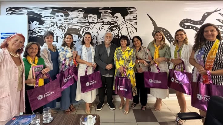 Delegation members holding purple SEBRAE Delas tote bags in front of a black and white woodcut mural.